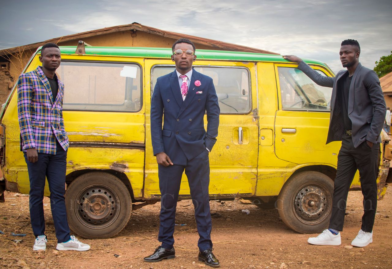 digital Three fashionable men in suits pose by a rustic yellow van in an outdoor setting.